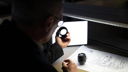 A man inspects a camera lens, technical drawings lie on the table.