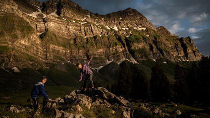 Two children climbing rocks in a mountain landscape at sunset.