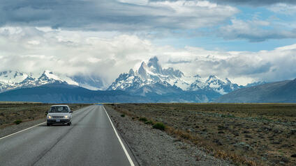 A car is driving on a road with snow-covered mountains in the background.