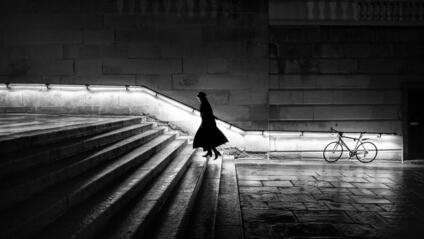 A woman in a long coat is walking up a staircase at night. The banister is illuminated.