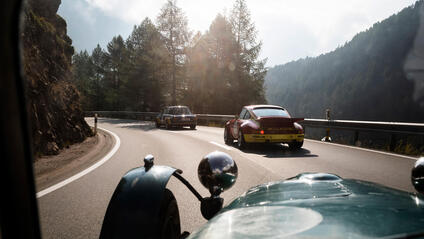 Vintage cars drive along a winding mountain road surrounded by trees and rocks.