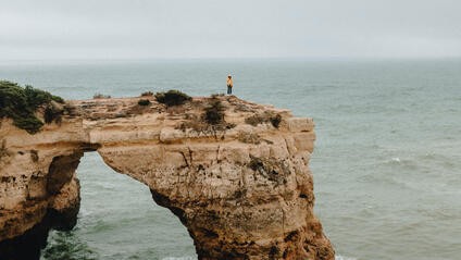 A person is standing alone on a rocky cliff jutting out into the ocean, and it is raining.