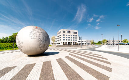 Large stone globe on a square in front of a hotel and parking lot under a blue sky.