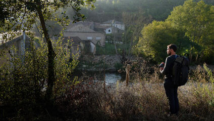 A man with a rucksack stands in the countryside and looks out over a small settlement by the river