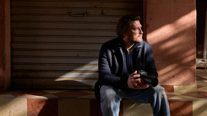 A man sits on a step with a camera in his hands, his face in the sunlight, in front of a closed shutter.