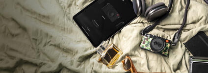 A camera, tablet, headphones, sunglasses, perfume and wallet laying on a bedcover.