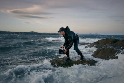 A person is standing on a rock in a stormy sea, holding a pair of binoculars.