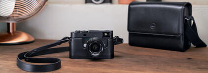 Leica camera on table with black leather case and fan in background.