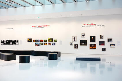 Exhibition of photos and names of photographers on a white wall, with black seats in front of it.