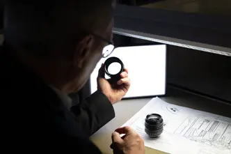 A man inspects a camera lens, technical drawings lie on the table.