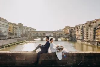 A wedding couple sits on a wall overlooking the river, surrounded by old buildings.