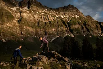 Two children climbing rocks in a mountain landscape at sunset.