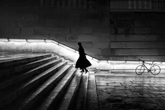 A woman in a long coat is walking up a staircase at night. The banister is illuminated.