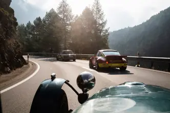 Vintage cars drive along a winding mountain road surrounded by trees and rocks.