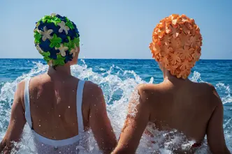 Two people in swimsuits and colorful bathing caps sitting in splashing water in the sea.
