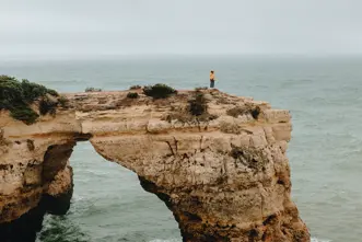 A person is standing alone on a rocky cliff jutting out into the ocean, and it is raining.