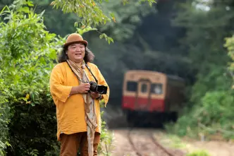 Man with camera stands beside railroad tracks and a train in background.