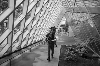 A cameraman walking with a professional camera setup in an architectural space.