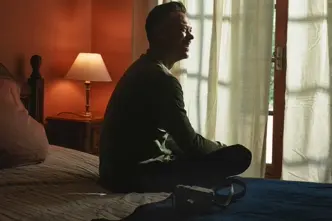 Smiling person sitting on a bed and a camera lying next to it.