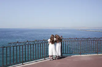 Two girls in white dresses stand at a railing and look to the sea.