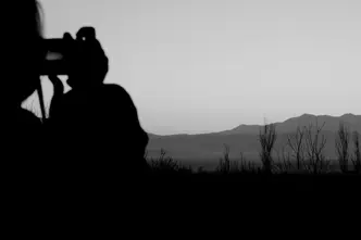 Black-and-white photograph of a person taking a picture of a lake and mountains in the background.