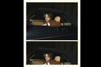 A person in a brown jacket sits in a car and looks through the open window.