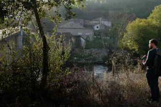 A man with a rucksack stands in the countryside and looks out over a small settlement by the river