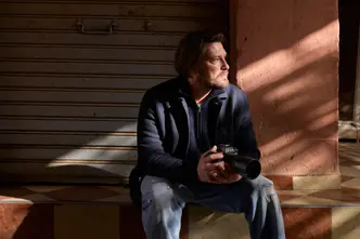 A man sits on a step with a camera in his hands, his face in the sunlight, in front of a closed shutter.