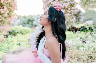 Woman in a pink dress and white gloves is sitting in a rose garden looking to the side.