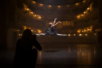 A person in the shade takes a picture of a ballerina jumping in an illuminated theatre.