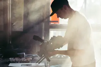 Person holds camera over a table with art materials, warm sunlight falls through window.