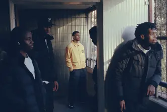 Five men in a shady hallway looking outside, the one in the middle wearing a yellow jacket.