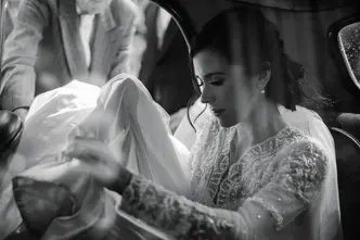 A bride sitting in a car holding her veil and a man standing next to the open car door.