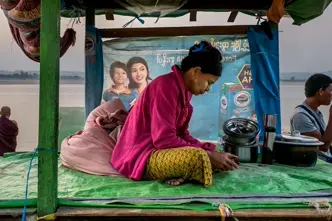 Woman sits on plastic tarp looking at a phone, in background are other people and a river.