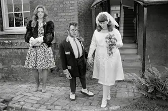 A wedding couple and a woman in a dress stand in front of a building.
