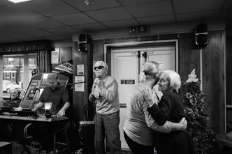 A man sings into a microphone while two older women dance and a man plays the keyboard in the background.