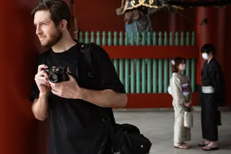 A photographer holds a camera while standing near traditional architecture, with two people in kimonos in the background.