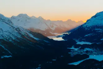 Snow-covered alpine valley at sunrise, with mountain peaks and lakes glowing in the morning light.