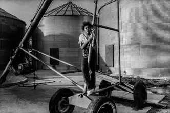 Young girl standing beside farm equipment and grain silos on a family farm in Arkansas, captured in black and white. A documentary photograph reflecting rural life, agricultural heritage, and generational connection.