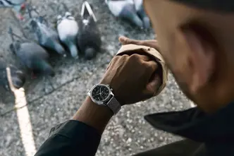 Jason Roman checks his Leica Watch while feeding pigeons in an urban public space, capturing a candid moment of everyday city life and human interaction with wildlife.