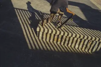 Skateboarder carrying a board through dramatic striped shadows at a skatepark, capturing movement, urban culture and graphic light.