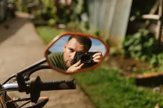 Photographer Philipp Gladsome reflected in a motorcycle mirror while taking a picture with a Leica M camera, surrounded by greenery in warm natural light.