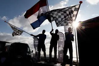 Silhouetted fans wave Dutch and chequered flags at a Formula 1 event in Zandvoort, with bright sunlight breaking through above the seaside circuit.