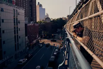Paul Hepper leaning through the fence of a New York City bridge, taking a street photograph above traffic and urban buildings in warm evening light.