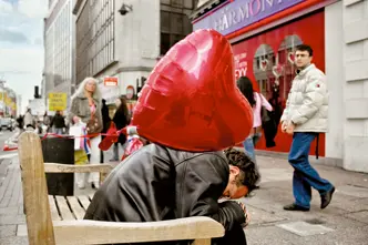 Street scene with a person seated on a bench, partially obscured by a large red heart-shaped balloon, surrounded by pedestrians in an urban setting.