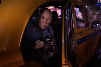 Photographer holding a Leica SL camera while seated inside a yellow taxi at night, illuminated by colorful urban lights.