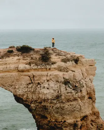 A person is standing alone on a rocky cliff jutting out into the ocean, and it is raining.
