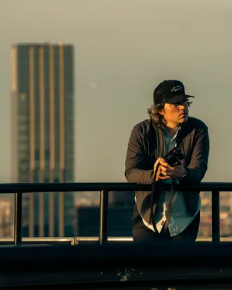 A man holding a camera leans against a railing with skyscrapers in the background.