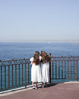 Two girls in white dresses stand at a railing and look to the sea.