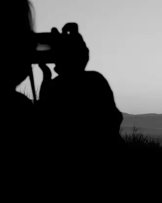 Black-and-white photograph of a person taking a picture of a lake and mountains in the background.